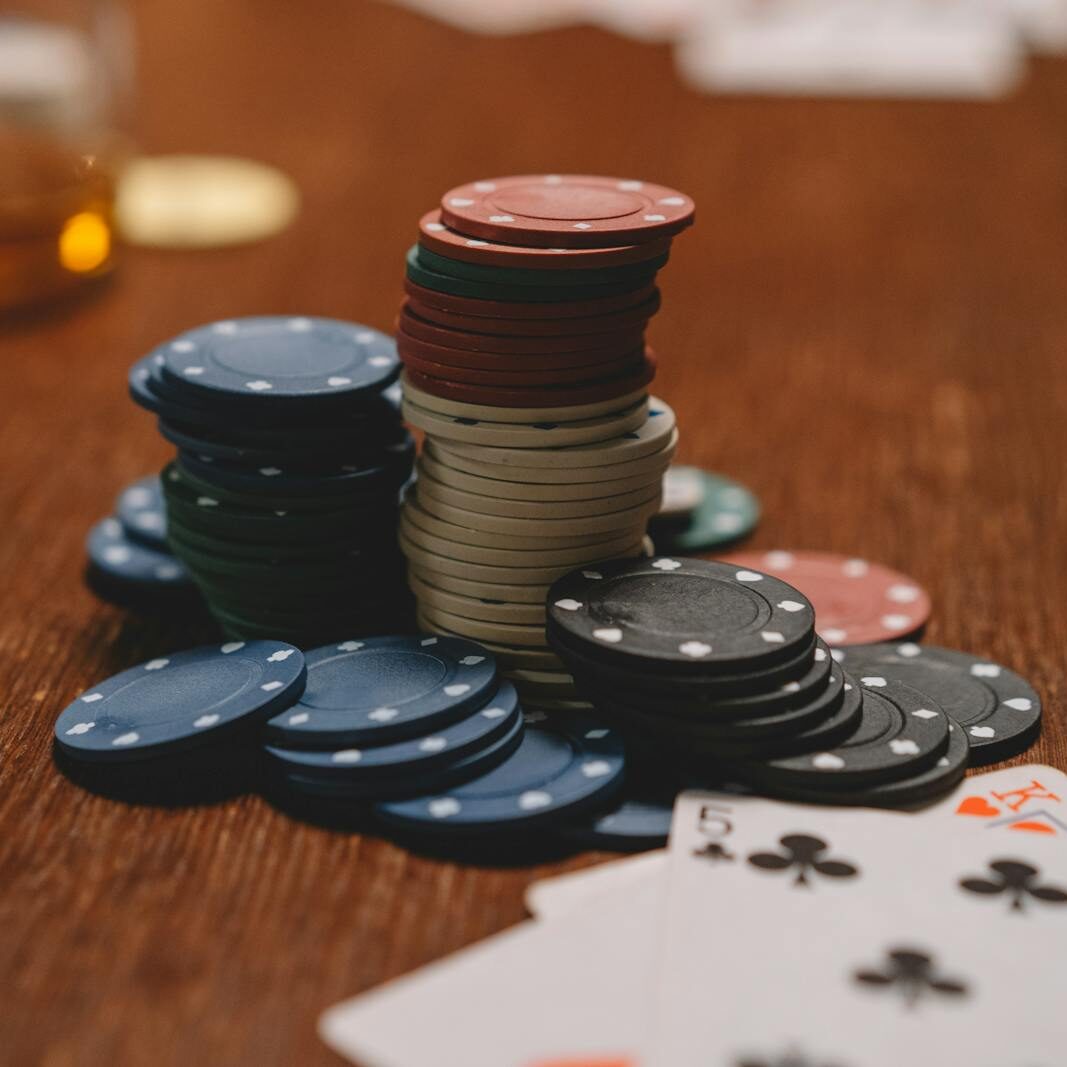 Poker chips stacked on a wooden table with playing cards and blurred glass in the background.